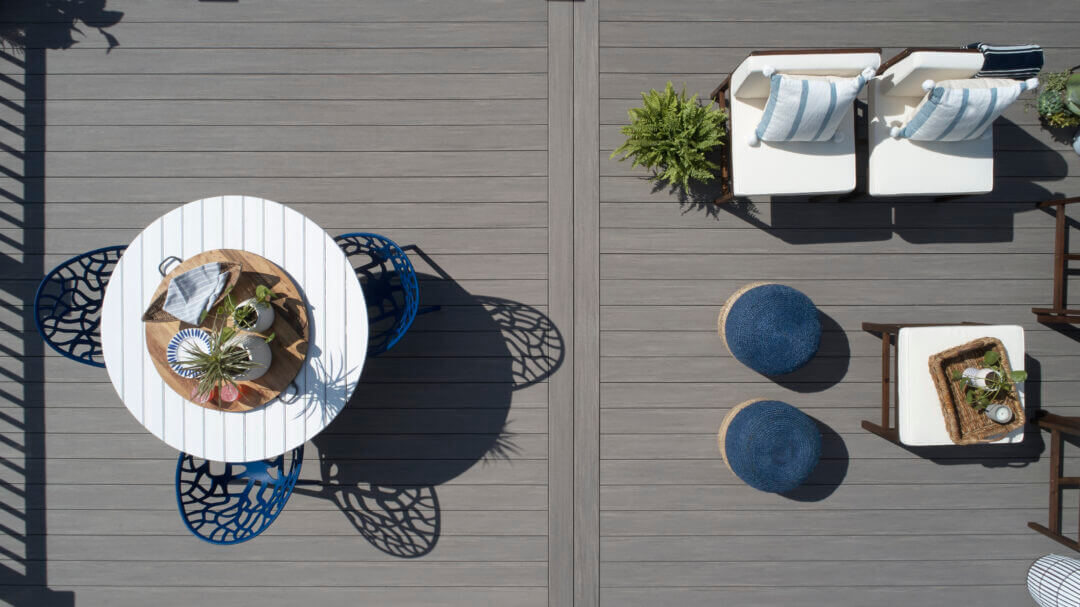 An overhead aerial view of a composite deck. The boards are gray and two splice boards run down the middle of the deck, cutting the deck in half. A white round table with three blue chairs around it sits on one side of the deck. An outdoor living room area on the other side of the deck features two matching chairs and two blue wicker poufs around an ottoman. The composite deck is made of Coastline boards from the TimberTech AZEK Vintage Collection.